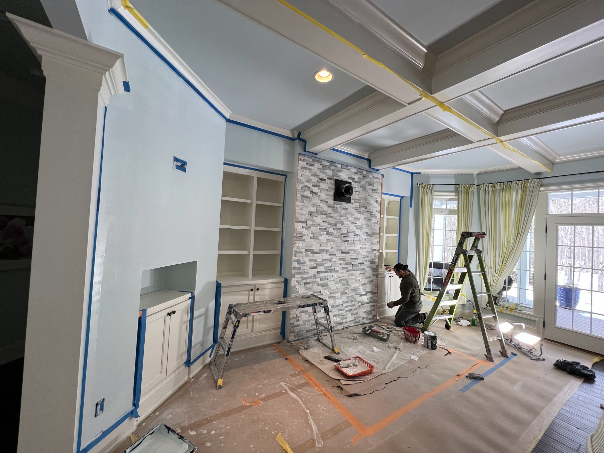 Interior living room with coffered ceiling, built-in shelves, stone fireplace accent, and blue painters tape during painting